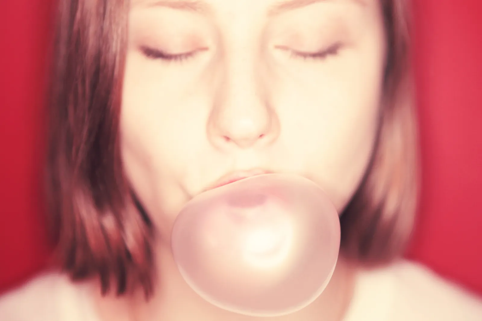 Person with closed eyes blowing a pale pink bubble against a red background