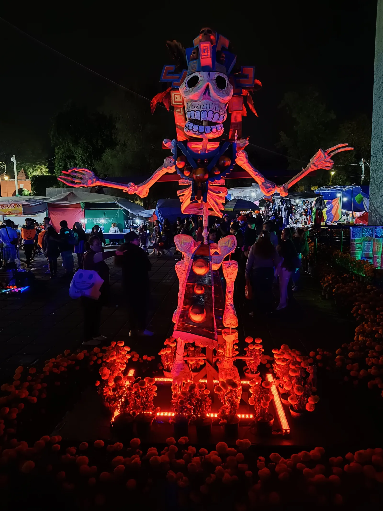An illuminated skeleton sculpture at a night-time Día de Muertos display, surrounded by orange flowers and visitors
