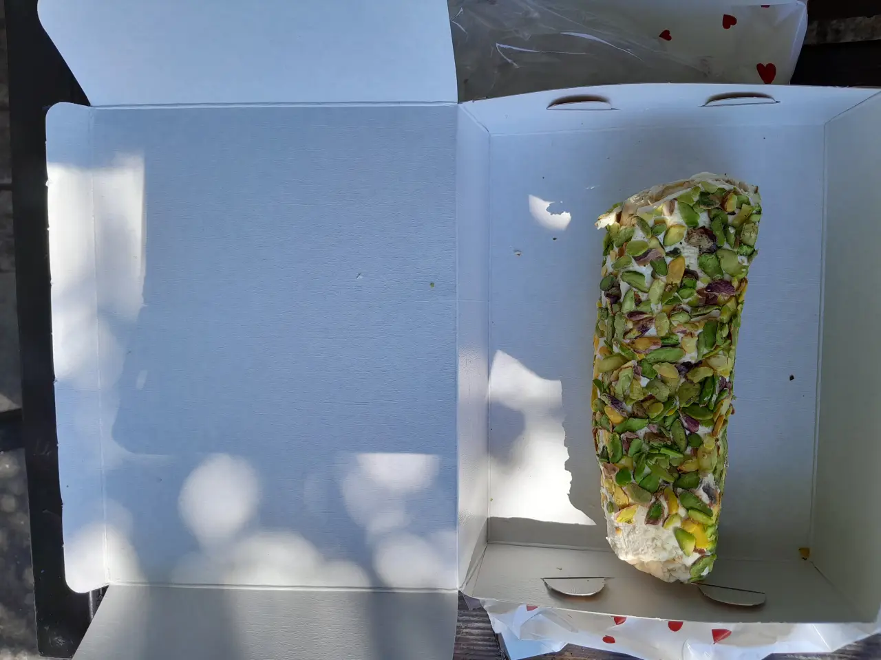 A long piece of Turkish delight covered in chopped green pistachios inside a white pastry box